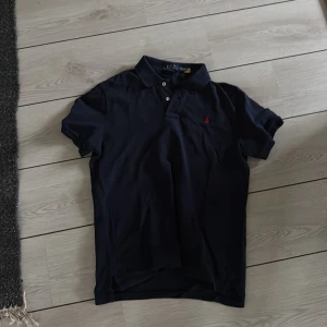 Mörkblå pikétröja från Ralph Lauren - Snygg mörkblå pikétröja från Ralph Lauren i slim fit-modell. Klassisk krage, korta ärmar och knäppning framtill. Ikoniska röda loggan broderad på bröstet. Perfekt för en clean och stilren look.Knappt använd