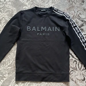 Svart Balmain tröja - Snygg svart sweatshirt från Balmain i storlek XS. Tröjan har Balmain Paris-tryck framtill och logga längs ena ärmen. Klassisk rund halsringning och långa ärmar. Perfekt för dig som vill ha en stilren och trendig look.