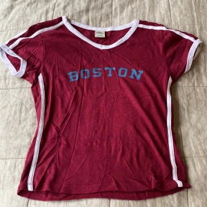 Röd Boston t-shirt från Urban Outfitters - Snygg röd t-shirt från Urban Outfitters med blått 'BOSTON'-tryck på bröstet. T-shirten har vita kontrastdetaljer längs ärmar och halsringning samt en sportig v-ringad design. Perfekt för en avslappnad och cool stil.