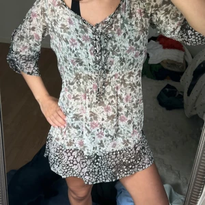 Blommig kortklänning med snörning - Söt kortklänning i transparent, lätt material med blommigt mönster i vitt, rosa och grått. Klänningen har trekvartsärm, snörning framtill och volangkant nertill. Perfekt för sommarens varma dagar och ger en romantisk vibe.