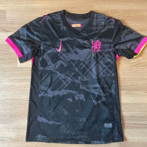 Chelsea Madueke 11 Nike fotbollströja - Chelsea fotbollströja med Madueke 11 på ryggen. Svart bas med grått mönster, rosa och gula detaljer på ärmarna samt Nike-logga i rosa/gult. Broderad Chelsea-lejon på bröstet. Tillverkad i lätt och ventilerande material, perfekt för match eller träning.