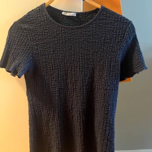 Marinblå ribbad t-shirt från Zara - Säljer en svart t-shirt från Zara i storlek M. T-shirten har korta ärmar och är gjord i ett ribbat, strukturerat material som ger en cool och modern känsla. Perfekt till jeans eller kjol för en avslappnad men trendig look.