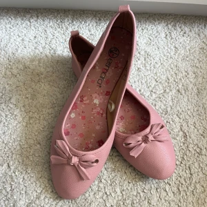 Rosa ballerinaskor med rosett från Esmara - Söta ballerinaskor i ljusrosa från Esmara med rund tå och dekorativ rosett framtill. Skorna har en lätt texturerad yta i lädermaterial och blommigt mönstrad innersula. Perfekta för dig som gillar en feminin och enkel stil. Innersulan är 24cm. Är borta just nu så kan skicka på måndag!