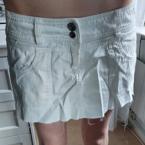 Vit jeanskjol med rå kant och dragkedjor - Vit kort jeanskjol med rå, fransig kant nertill. Två stora fickor fram och bak, bakfickorna har snygga broderier och dragkedjor med ringdetalj.  Ena fickan saknar en del av dragkedjan, kolla bild. Kjolen har dubbla knappar i midjan och bälteshällor. Perfekt för en cool och avslappnad stil. Storlek M/38