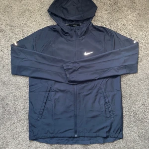 Nike Miller navy blue - Snygg marinblå vindjacka från Nike med huva och dragkedja framtill. Jackan har två sidofickor och reflekterande detaljer på ärmarna samt klassisk Nike-logga på bröstet. Perfekt för löpning eller när du vill ha en lätt jacka med sportig vibe.