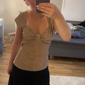 Glittrig beige topp från Bubbleroom - Supersnygg glittrig beige topp från Bubbleroom med korta ärmar och en snygg twistad detalj framtill vid bysten. Toppen har en figurnära passform och är perfekt när du vill sticka ut lite extra. Materialet är mjukt och stretchigt för en bekväm känsla.