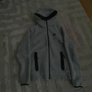 Grå Nike zip-hoodie med huva - Snygg grå zip-hoodie från Nike med svart logga på bröstet och svarta dragkedjor på fickorna. Jackan har huva, hel dragkedja och sportig look. Perfekt för chill eller träning, tillverkad i mjukt material som känns skönt mot huden.