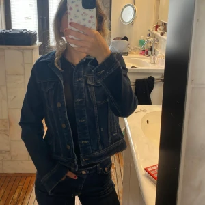 Mörkblå jeansjacka med knappar - Snygg mörkblå jeansjacka med klassisk passform och tydliga kontrastsömmar. Jackan har bröstfickor med lock och metallknappar framtill. Perfekt för dig som gillar en tidlös och cool look.