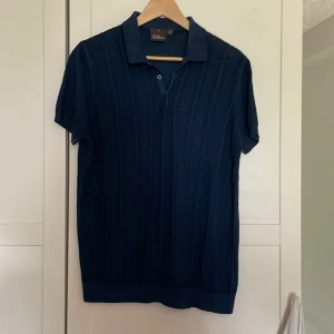 Mörkblå pikétröja Oscar Jacobson M - Snygg mörkblå pikétröja från Oscar Jacobson, tillverkad i Italien. Tröjan har korta ärmar, klassisk krage och knäppning framtill. Använd ett fåtal gånger, mycket bra skick. 