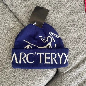 Blå mössa från Arc'teryx - Snygg blå mössa från Arc'teryx med vit logga och fossilmotiv framtill. Klassisk uppvikt kant och stickad i mjukt material som håller dig varm. Perfekt för kalla dagar och streetstyle. Nypris runt 600