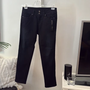Svarta jeans från Fransa Jeans, strl 38 - Svarta jeans från Fransa Jeans med snygga detaljer som dubbla knappar i midjan och dragkedja på fickan. Klassisk femficksmodell i stretchig bomullsblandning som sitter skönt och har en clean look. Perfekta till sneakers eller boots.