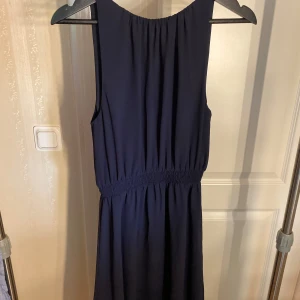 Mörkblå/svart  ärmlös klänning med resår - Mörkblå/svart klänning med ärmlös design och rund halsringning. Klänningen har resår i midjan för en skön passform och ett lätt, flygigt tyg som ger en fin siluett. Perfekt för dig som gillar stilrena och enkla plagg med en elegant touch.