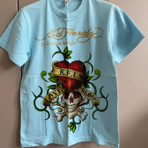 Ljusblå  t-shirt med tryck - Snygg ljusblå t-shirt  med stort färgglatt tryck framtill – dödskalle, hjärta och banderoll med texten 'Love Kills Slow'. Guldigt logotryck både fram och bak. Klassisk passform och korta ärmar. Perfekt statement-plagg för dig som gillar streetstyle.