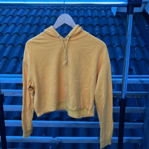 Gul croppad hoodie från H&M - Gul hoodie från H&M Basic i croppad modell med huva och snörning. Tröjan har långa ärmar och är gjord i mjuk bomullsmix, perfekt för en chill och avslappnad stil. Passformen är normal och tyget är skönt mot huden.