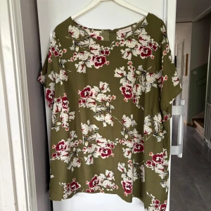 Olivgrön blommig blus med dragkedja - Olivgrön blus från Zizzi med stora vita och vinröda blommor. Blusen har korta ärmar, rund halsringning och en diskret dragkedja i ryggen. Luftig och skön passform, perfekt för dig som gillar färg och mönster.