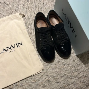 Svarta sneakers från Lanvin i mocka - Snygga svarta sneakers från Lanvin med ovandel i mocka och lackad tå. Skorna har rund tå, svarta snören och diskreta metalldetaljer vid snörningen. Klassisk och stilren modell som passar perfekt till streetwear eller en mer dressad look. Uk 9/ 43-44 i bra skick pris kan diskuteras