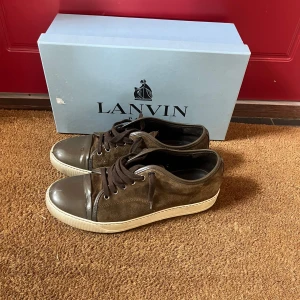 Bruna sneakers från Lanvin i mocka - Snygga bruna sneakers från Lanvin med ovandel i mocka och lackad tå samt häl i mörkbrunt. Skorna har vita platta sulor och bruna snören. Perfekta för dig som gillar stilrena och exklusiva sneakers med en twist. Äkthetscetrifikat finns och tas privat.