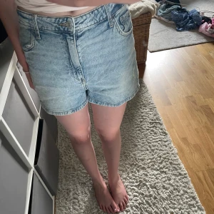 Ljusblå högmidjade jeansshorts - Säljer ett par ljusblå jeansshorts med klassisk femficksmodell och hög midja. Shortsen har en avslappnad passform och är tillverkade i mjukt denimtyg. Perfekta för varma dagar och enkla att matcha med allt. Dragkedja och knapp framtill.