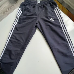 Mörk blå Adidas byxor med vita ränder - Säljer ett par mörk blåa Adidas byxor med klassiska vita ränder längs sidorna och broderad logga framtill. Byxorna har elastisk midja, sidfickor och är gjorda i ett glansigt polyestermaterial. Perfekta för en sportig och avslappnad stil.