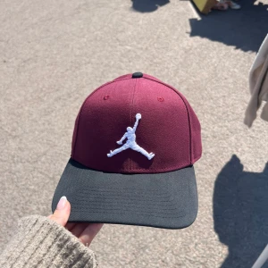 Vinröd och svart Jordan keps - Snygg keps från Jordan i vinrött med svart skärm och vit broderad Jumpman-logga framtill. Klassisk form med böjd skärm och ventilerande hål upptill. Perfekt accessoar för en sportig och trendig look.