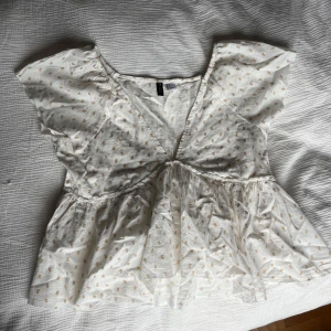 Vit blommig blus från H&M - Supersöt vit blus från H&M Divided med små beige blommor, v-ringning och volang nedtill. Luftigt tyg i bomull och korta ärmar. Perfekt för vår och sommar.