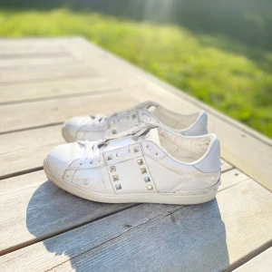 Valentino Rockstud vita sneakers - Snygga vita Valentino Rockstud sneakers i skinn med ikoniska nitar i guld längs sidorna och runt hälen. Klassisk rund tå, vita snören och platt sula. Perfekta för dig som vill ha en lyxig och edgy touch till din outfit. Storlek 41.5 passar 42.5  kan sänka pris vid snabbköp