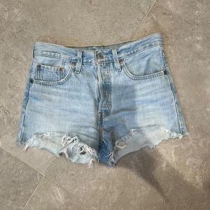 Levi's ljusblå jeansshorts med fransar - Snygga ljusblå jeansshorts från Levi's med klassisk femficksdesign och råa fransiga kanter. Shortsen har hög midja och den ikoniska Levi's-lappen bak. Perfekta för sommaren och ger en avslappnad vibe.