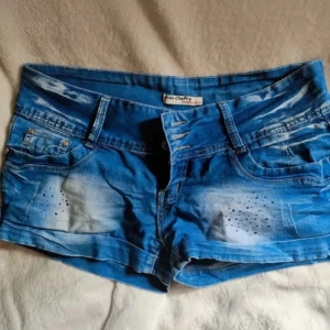 Blå jeansshorts från Miss Cherry - Snygga blå jeansshorts från Miss Cherry med slitna detaljer och coola nitar framtill. Shortsen har flera knappar i gylfen och klassiska fickor. Perfekta för sommaren när du vill ha en avslappnad och trendig look.