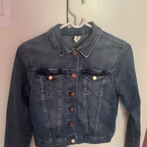 Klassisk blå jeansjacka från H&M - Snygg blå jeansjacka från H&M med klassisk krage, kopparfärgade knappar och två bröstfickor med lock. Jackan har en rak passform och är tillverkad i stretchig denimtyg. Perfekt att slänga över en huvtröja eller t-shirt för en cool eller casual look.