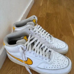 Nike Air Force 1 High vit/gul - Nike Air Force 1 High sneakers i vitt skinn med gula och lila detaljer. Klassisk hög modell med snörning, perforerad tå och vadderad plös. Swoosh-logga i gult med lila kant. Perfekta för dig som gillar retrovibbar och streetstyle.