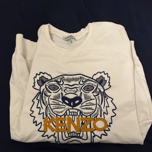Vit tröja med tigertryck från Kenzo - Säljer en stilren vit tröja från Kenzo med ett ikoniskt tigertryck i blått och orange på framsidan. Tröjan har långa ärmar och en rund halsringning. Perfekt för dig som vill ha en trendig och bekväm look.