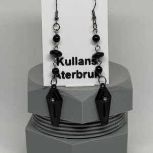 Svarta örhängen från Kullans Återbruk - Unika svarta örhängen med en cool design från Kullans Återbruk. De har svarta pärlor och en kistformad hängande detalj. Perfekta för att ge en edgy touch till din stil.