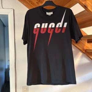 Svart t-shirt från Gucci - Snygg svart t-shirt från Gucci i storlek L (oversized) med ett coolt rött och vitt tryck på framsidan. Perfekt för att ge din outfit en lyxig touch. Tillverkad i Italien.