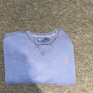 Blå tröja från Polo Ralph Lauren - Säljer en snygg blå tröja från Polo Ralph Lauren med det klassiska röda logotypen på bröstet. Tröjan är tillverkad i en bomullsblandning och har långa ärmar med ribbade muddar. Perfekt för en stilren och avslappnad look.