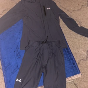 Blå tracksuit  från Under Armour - Säljer en blå tracksuit från Under Armour med dragkedja och huva. Tracksuiten har en diskret logga på bröstet och en ficka med dragkedja. Perfekt för utomhusaktiviteter.