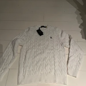 Vit stickad tröja från Ralph Lauren - Säljer en stilren vit stickad tröja från Ralph Lauren med kabelstickat mönster. Tröjan har långa ärmar och en liten broderad logotyp på bröstet. Perfekt för kyligare dagar.