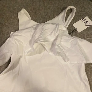 Vit blus från Zara - Säljer en stilren vit blus från Zara med volangdetaljer framtill. Blusen har breda axelband och volanger framtill verkligen perfekt till våren så gullig💕 pris kan diskuteras kontakta för fler bilder !!