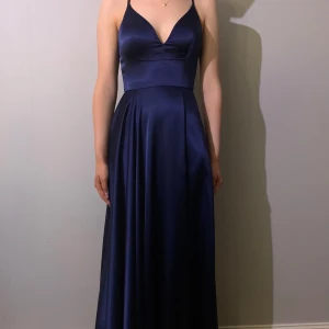 Marinblå balklänning från Ivoryfield stl 34 - Elegant marinblå långklänning i satin med v-ringad urringning och smala axelband. Klänningen har en stilren slits och en vacker öppen rygg med snörning. Använd vid ett tillfälle och sedan kemtvättad. Modellen heter Stella och är upplagd för att passa en person som är 170 cm i klackar.