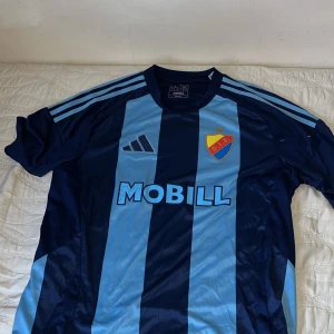 Blå och svart fotbollströja från Adidas - Snygg fotbollströja från Adidas i blått och svart med klubbmärke på bröstet. Tröjan har korta ärmar och klassiska ränder på axlarna. Perfekt för fotbollsfans!