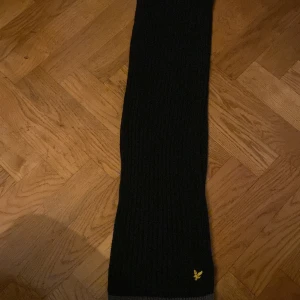Svart halsduk från Lyle & Scott - Snygg svart halsduk från Lyle & Scott med gråa detaljer i ändarna. Den har en diskret logga i gult som ger en stilren touch. Perfekt för att hålla värmen under kyliga dagar.