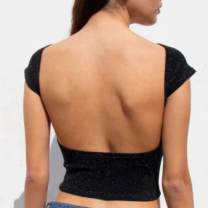 Svart glittrig topp med öppen rygg subdued  - Snygg svart glittrig topp med en djärv öppen rygg. Toppen har korta ärmar och en tight passform som framhäver figuren. Perfekt för en kväll ute eller en festlig tillställning. Storlek M men passar xs-m