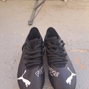Svarta fotbollsskor från Puma - Snygga svarta fotbollsskor från Puma med vit logga och text. Skorna har snörning för en säker passform och är designade för att ge bra grepp på planen. Perfekta för fotbollsträning och matcher. Använda ungefär 20 gånger då skorna blev för småa så fort. (Möts upp i Jönköping) tyvärr ingen box, tvättas innan den säljs