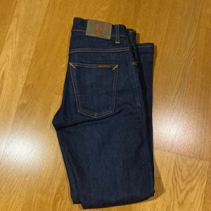 Mörkblå jeans från Nudie Jeans - Snygga mörkblå jeans från Nudie Jeans med orange sömmar. De har en klassisk femficksdesign och en knappgylf. Perfekta för en stilren look.