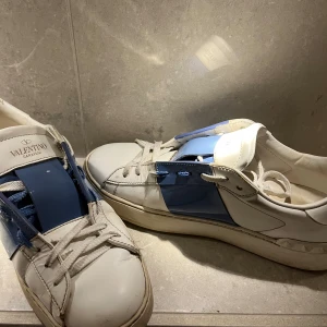 Valentino Garavani sneakers i vitt och blått - Snygga Valentino Garavani sneakers i vitt med blå detaljer. Skorna har en unik design med breda blå remmar över snörningen och är tillverkade i skinn. Perfekta för en stilren look. Jag tar också emot byten, storleken är 38,5 men passar 39,5