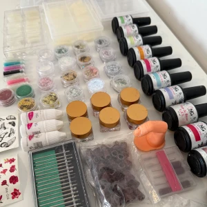 Nagelprodukter  - Ett set med olika nagelprodukter inklusive glitter, nagellack och dekorationer. Flera små burkar med glitter i olika färger, flaskor med nagellack i olika nyanser och små dekorationer för naglar. Perfekt för att skapa kreativa nageldesigner hemma.