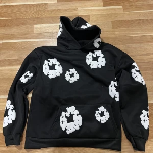 Denim tears - En riktig bra hoodie inför våren. En svart hoodie från Denim Tears med unikt vitt mönster. Tröjan har en stor ficka framtill och en bekväm huva. Perfekt för en cool och avslappnad stil.