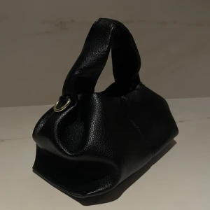 Svart handväska med axelrem - Snygg svart handväska i skinn imitation med en avtagbar axelrem. Väskan har en stilren design med guld detaljer. Perfekt för att bära både i handen och över axeln.