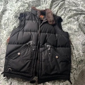 Svart dunväst från Parajumpers - Säljer en svart dunväst från Parajumpers med dragkedja och bruna detaljer. Västen har en quiltad design och är perfekt för kyligare väder. Den har praktiska fickor med dragkedjor och en mjuk krage.