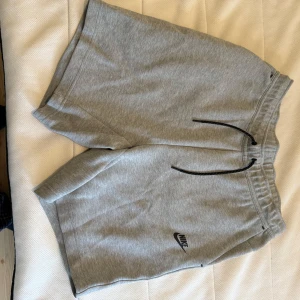 Grå shorts från Nike - Säljer ett par grå shorts från Nike med elastisk midja och snörning. De har en avslappnad passform och praktiska fickor med dragkedja. Perfekta för en bekväm och sportig stil.
