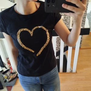 Svart t-shirt med leopardhjärta - Säljer en svart t-shirt med ett stort leopardmönstrat hjärta på framsidan. T-shirten har korta ärmar och en rund halsringning. Perfekt för en avslappnad stil med en touch av djurmönster.